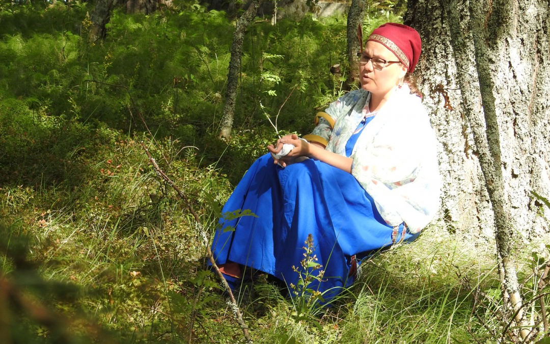 Itkuvirsien itkijä Emilia Kallonen istuu puun juurella karjalaisnaisen asussa